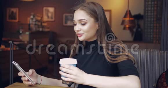 年轻漂亮的女孩坐在咖啡馆的桌子旁手里拿着一杯咖啡浏览着智能手机视频的预览图