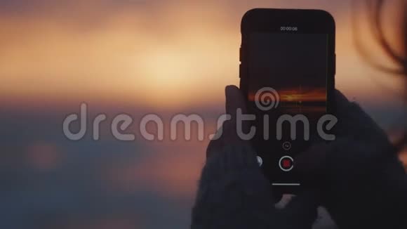 特写快乐旅游女人拍摄智能手机视频史诗粉红色日落天空风景在风海海岸慢动作视频的预览图