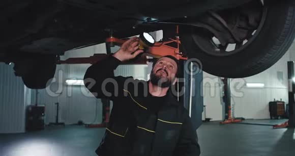 在汽车车库里一个穿制服的成熟的机械师站在车下一边用一个视频的预览图