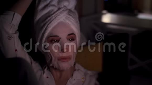 年轻女子戴着化妆面料面具头上戴着白色毛巾手里拿着智能手机听着看着晚上躺着视频的预览图