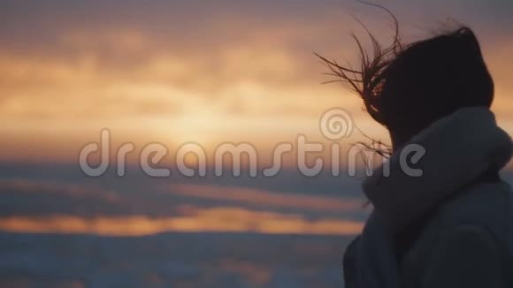 后景快乐的旅游女人头发在大风中吹着看着史诗般的粉红色日落在寒冷的海边慢动作视频的预览图