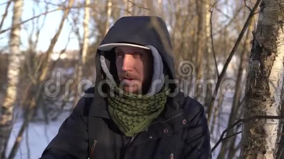 一个戴着帽子围巾和带罩的蓝色夹克的人站在森林里看着摄像机留胡子的肖像视频的预览图