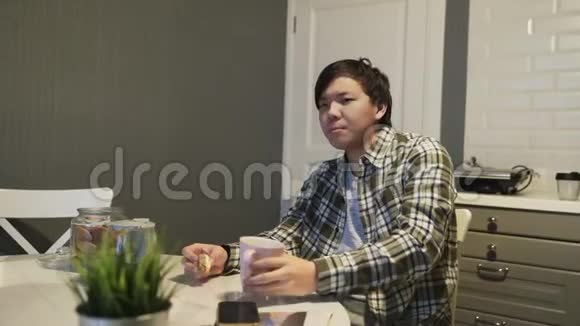 困乏的亚洲男人在桌旁喝咖啡吃饼干视频的预览图