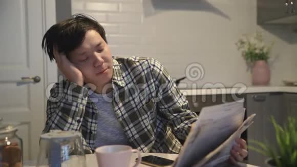 昏昏欲睡的亚洲男人在桌旁喝咖啡看报纸视频的预览图