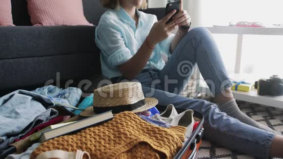 一个年轻女孩在旅行箱附近看着智能手机上的社交网络和朋友聊天视频的预览图