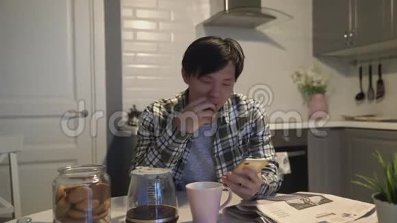 快乐的亚洲男人在智能手机上读一些有趣的东西在家厨房里微笑视频的预览图