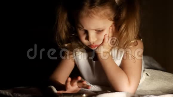 小女孩躺在床上在黑暗中玩智能手机孩子的近脸被明亮的灯光照亮视频的预览图