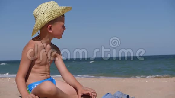 男孩坐在金沙上这孩子戴着一顶大帽檐的草帽孩子在沙滩上玩视频的预览图