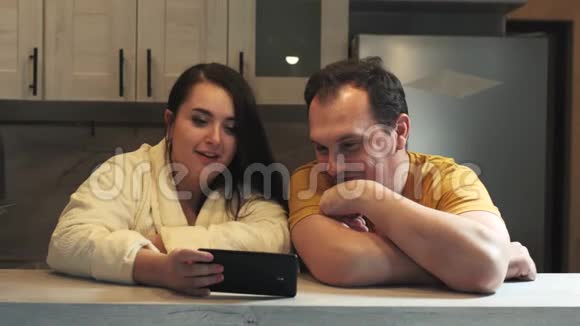 一对幸福的家庭夫妇坐在厨房的木桌旁使用智能手机分享应用程序或联系人视频的预览图