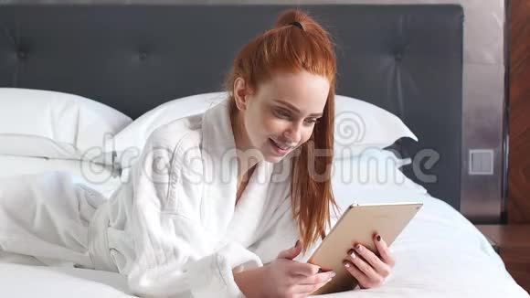 年轻的红头发妇女使用平板电脑躺在酒店房间的床上视频的预览图