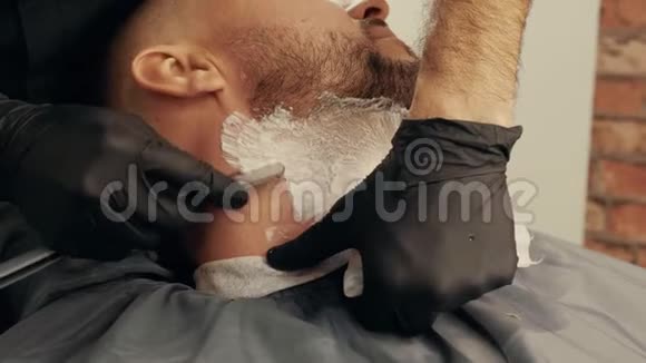 在男式沙龙里用直剃须刀把理发师的手刮胡子用剃须刀和泡沫刮胡子视频的预览图