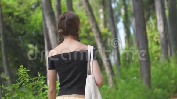 年轻女子拿着白色的袋子在绿林中漫步视频的预览图