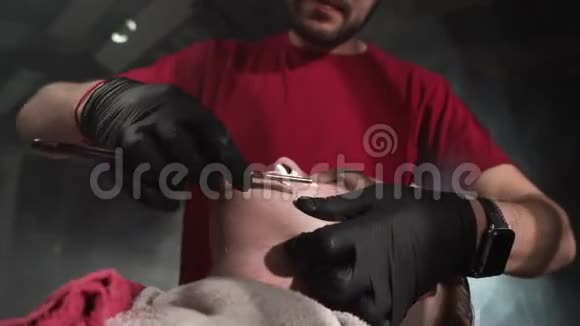 穿着红色T恤和黑色手套的理发师用危险的剃刀在理发店给顾客刮胡子视频的预览图