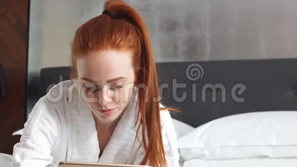 年轻的红头发妇女在酒店房间的床上躺着平板电脑看电影视频的预览图
