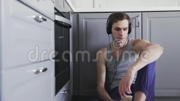 年轻人在智能手机上听音乐戴着耳机坐在厨房的地板上视频的预览图