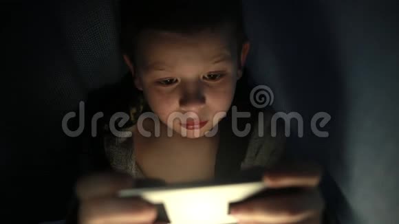 毯子下面的小家伙晚上在他的手机智能手机上玩游戏视频的预览图