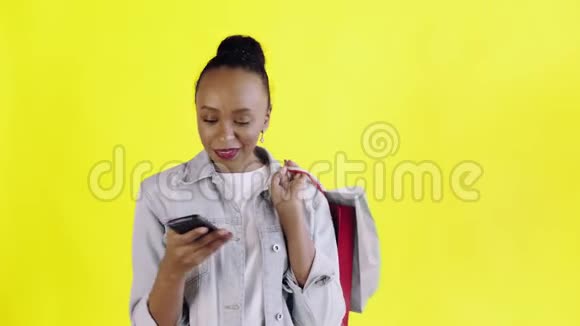 美国黑人妇女带着购物袋的画像正在黄色背景下通过智能手机录制语音信息视频的预览图
