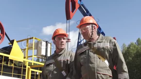 两名戴着橙色头盔的石油工人在油泵千斤顶附近行走和交谈石油工程师监督原油现场视频的预览图