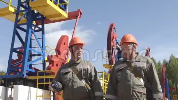 两名男性石油工人在油泵千斤顶附近行走和交谈石油工程师监督原油钻井平台视频的预览图
