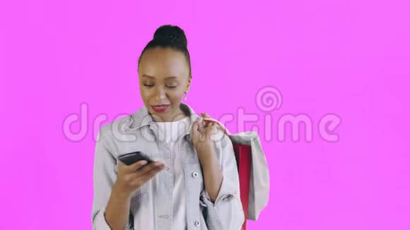 美国黑人妇女带着购物袋的肖像正在粉色背景上通过智能手机录制一条语音信息视频的预览图