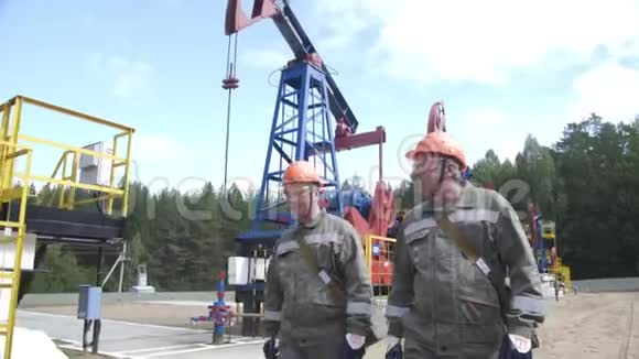 两名身穿橙色头盔的石油工人在油泵千斤顶和井口连接油设备附近行走和交谈视频的预览图