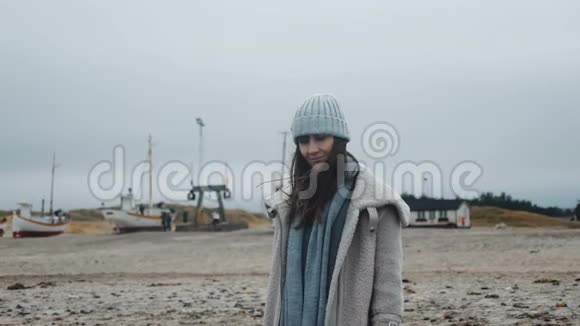 美丽的快乐女游客在冬季大衣在风低潮冷波罗的海海滩与船慢运动的肖像视频的预览图
