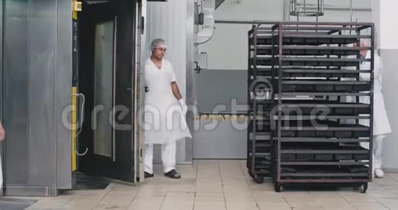 大的面包店里的工人穿着白色制服把货架上的生面包装在工业烤箱里以获得烘焙食品视频的预览图