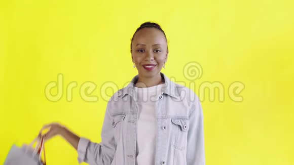 美国黑人妇女带着购物袋的肖像在工作室的黄色背景上竖起大拇指琼夹夹克视频的预览图