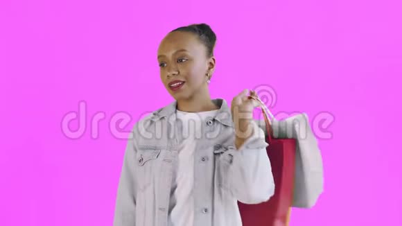 美国黑人妇女带着购物袋的肖像是在工作室的粉色背景上通过智能手机谈论的琼夹夹克视频的预览图
