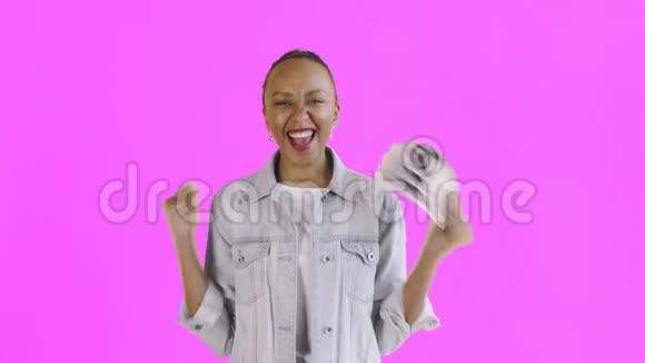 满意的兴奋的兴奋的非裔美国妇女在粉红色背景的Jean夹克上展示金钱和相机跳舞视频的预览图