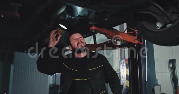 一个心情很好的机械师在车库里损坏的汽车下面用手电筒他试图找出问题所在视频的预览图
