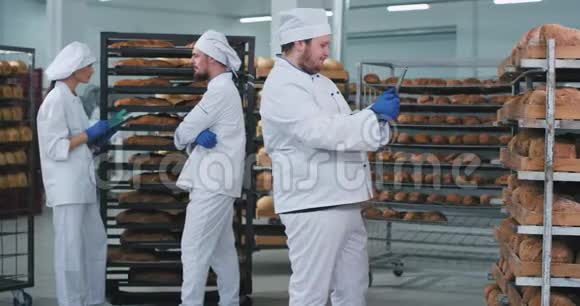 在一个大面包店里主面包师从货架上的一大片新鲜烘焙面包上拍照而另外两个人视频的预览图