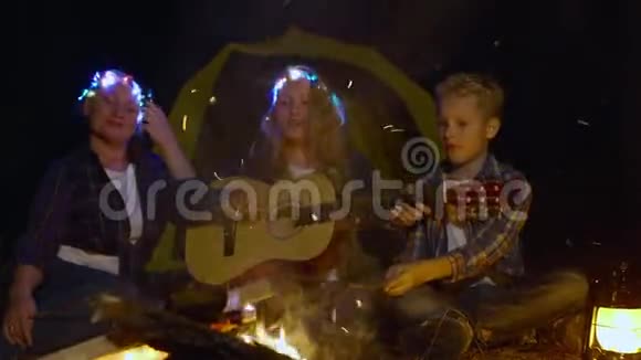 少女一起用吉他唱歌曲妈妈和哥哥在闪闪发光的篝火背景下女孩在演奏音乐视频的预览图