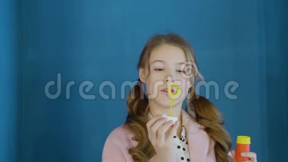 可爱的少年女孩吹着肥皂泡在蓝色的墙壁背景上微笑漂亮的女孩卷发视频的预览图
