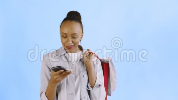 美国黑人妇女带着购物袋的肖像正在蓝色背景下通过智能手机录制语音信息视频的预览图