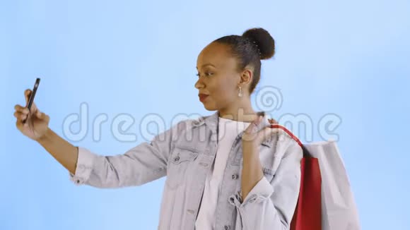 非洲裔美国妇女带购物袋的肖像正在工作室的蓝色背景上自拍琼夹夹克视频的预览图
