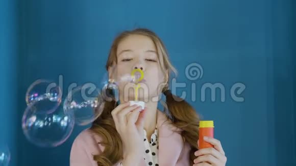 快乐女孩少年在蓝色工作室吹肥皂泡前相机两条辫子吹着的年轻姑娘视频的预览图