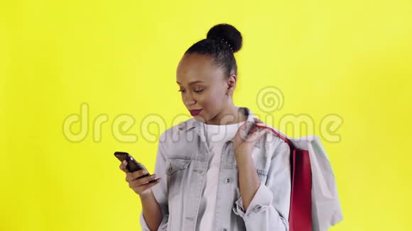 美国黑人妇女带着购物袋的肖像在工作室的黄色背景上通过智能手机交谈让视频的预览图