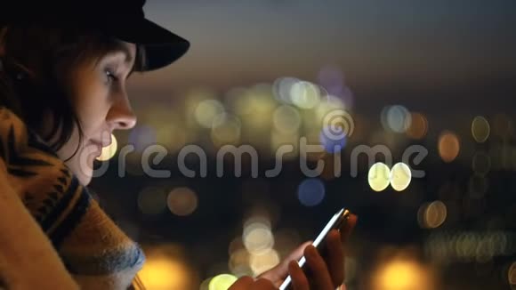 特写幸福美丽的女人在模糊的夜晚城市背景下使用智能手机她的脸被屏幕慢动作照亮视频的预览图