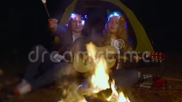头上戴着照明花环的女人在篝火旁享受吉他和烹饪棉花糖视频的预览图