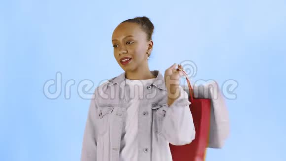 美国黑人妇女带着购物袋的肖像正在工作室的蓝色背景上通过智能手机交谈珍妮夹克视频的预览图