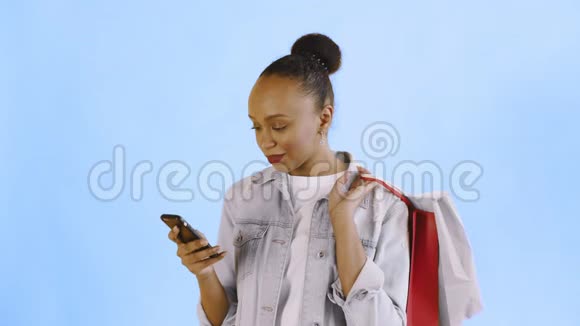 美国黑人妇女带着购物袋的肖像正在工作室的蓝色背景上通过智能手机交谈琼夹夹克视频的预览图