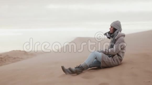 快乐的年轻白种人女游客摆姿势坐在寒冷的沙丘海滩下极端的风沙暴缓慢的运动视频的预览图