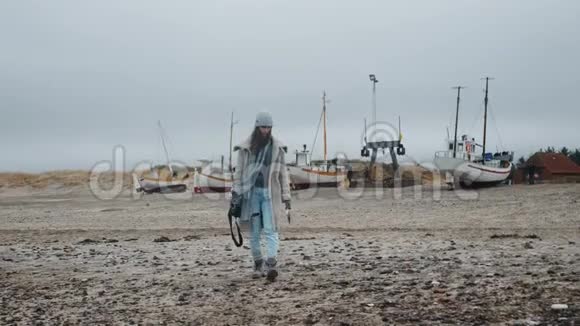 快乐的女摄影师穿着冬衣在干的低潮沙性波罗的海海滩上与停靠的船只慢动作漫步视频的预览图