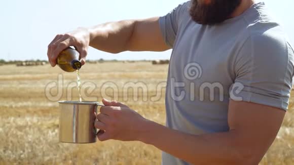 一个人把啤酒倒进麦田里的金属玻璃里上面有捆新鲜啤酒胡须男太阳田视频的预览图