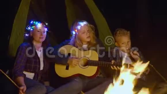 十几岁的女孩在森林远足中弹吉他唱着篝火前的歌友好的家庭在露营时唱歌视频的预览图