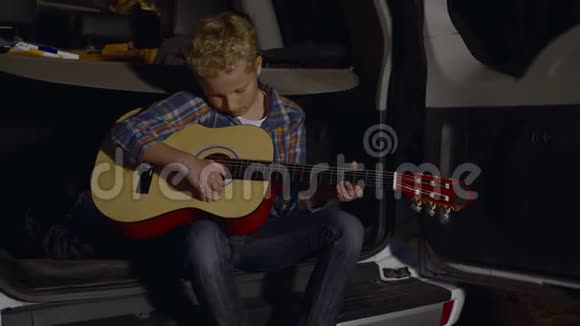 坐在汽车后备箱里用吉他演奏音乐的少年男孩旅游男孩吉他手在夏令营中演奏音乐视频的预览图