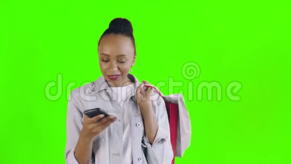 美国黑人妇女带着购物袋的肖像正在绿色背景下通过智能手机录制语音信息视频的预览图