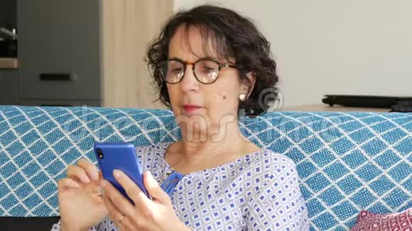 黑发开朗的高级女性坐在沙发上用智能手机视频的预览图