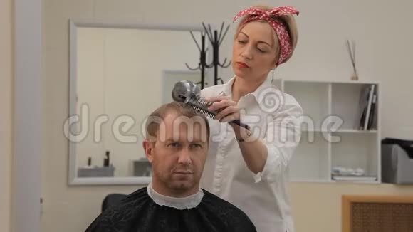 一个漂亮的理发师女孩用吹风机把头发擦干给一个男性客户她头发上有一个红色的头带视频的预览图